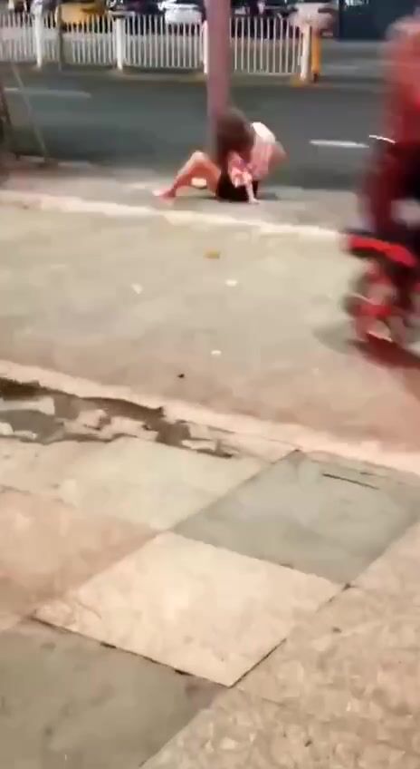 She masturbates against a tree in a public place, surrounded by people and cars.